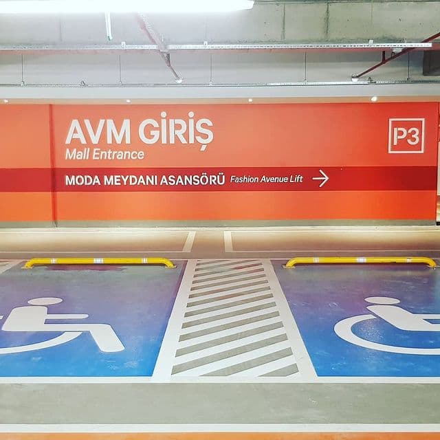AVM kapalı otoparkında mavi zeminli engelli park yerleri, aradaki beyaz tarama alanı ve sarı tekerlek stoperleri